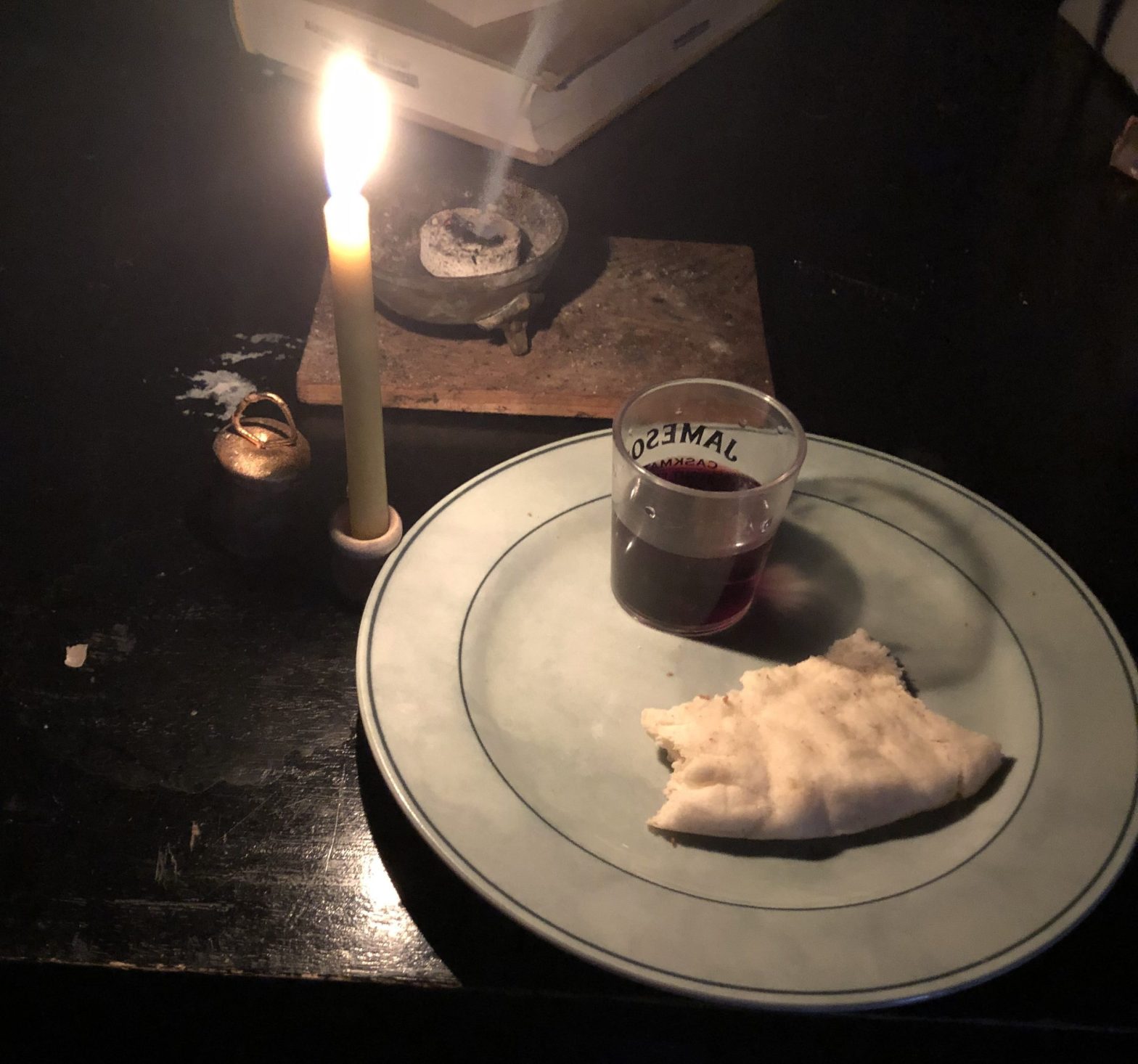 lighted candle next to green plate with shot glass of wine and pita bread. incense burning. evokes eucharistic table